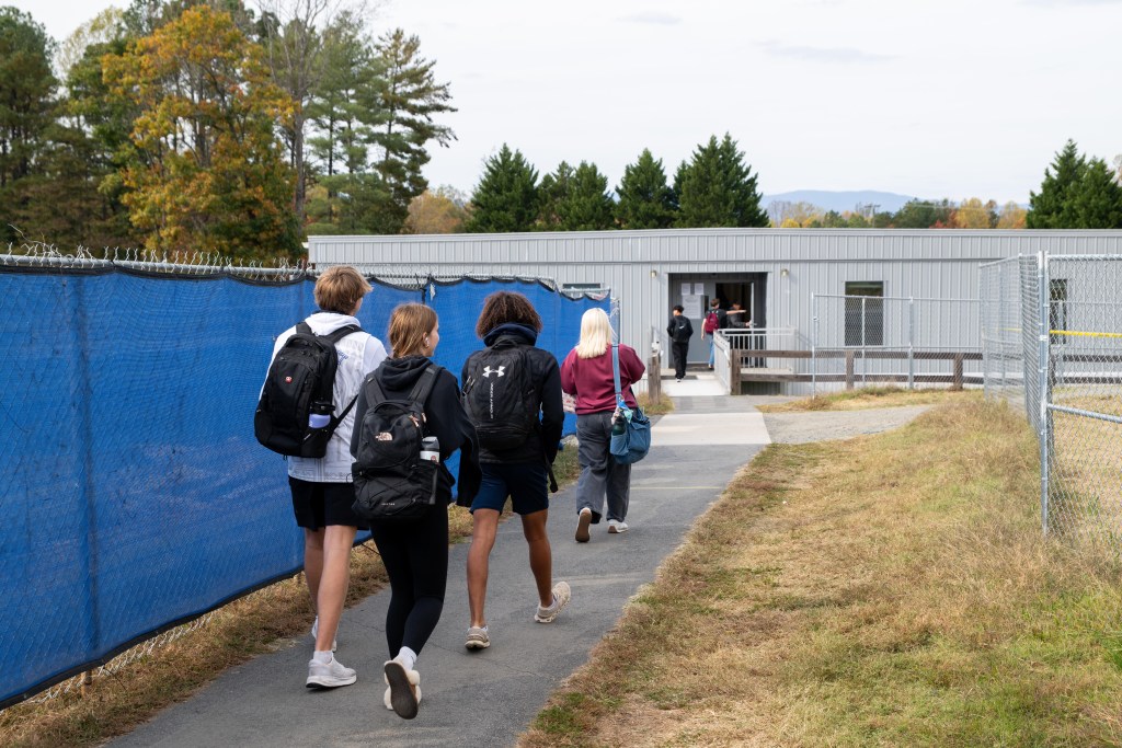 High school students wearing backpacks walk by a chain link fence with a blue tarp covering it, heading toward a long rectangular trailer with a door and small windows.