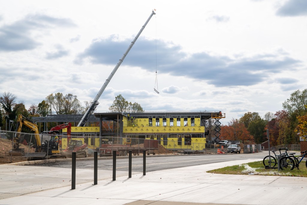 A digger and a large crane sit next to a building under construction. The crane is lifting a large girder above the building, and a chainlink fence closes off the construction site. Across the street, bikes are lined up on a small bike rack.