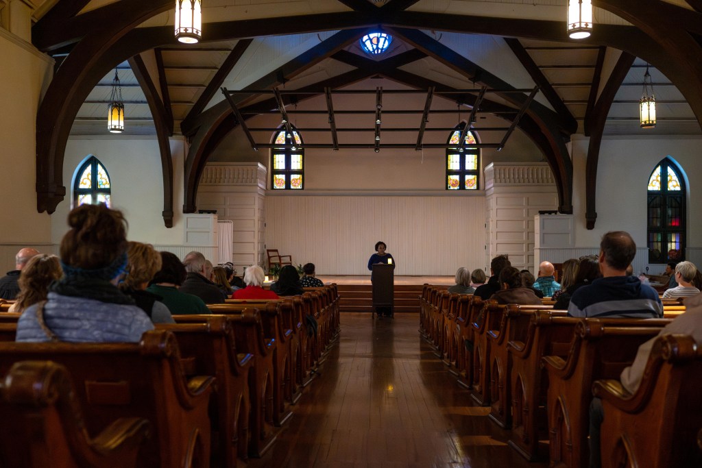 A photograph of a former church sanctuary with vaulted ceilings, old wooden pews, and stained glass. The image is symmetrical, taken looking down the center of an aisle dividing rows of pews. A few dozen people sit in the pews and listen to a woman standing at a lectern.