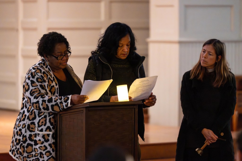 A photograph of three women. Two of the women stand at a lectern, a flickering candle between them. Each is looking at a piece of paper in her hands. A third woman wearing religious garb, stands to their right, holding a bell. All three look like they are grieving.