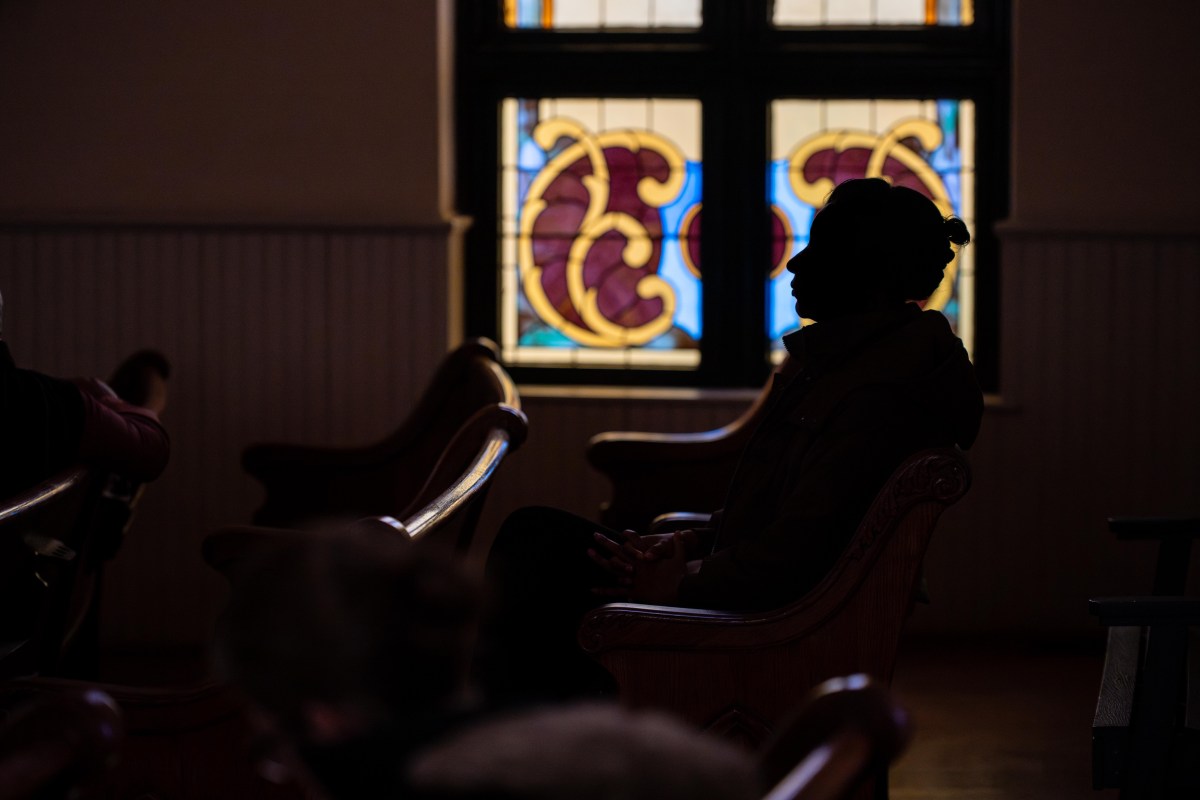 A photograph taken inside a former church sanctuary. A person sits in old wooden pews, and only her profile is visible. The rest of her face and body are a shadow. Behind her are stained glass windows softly illuminated with afternoon sunlight.