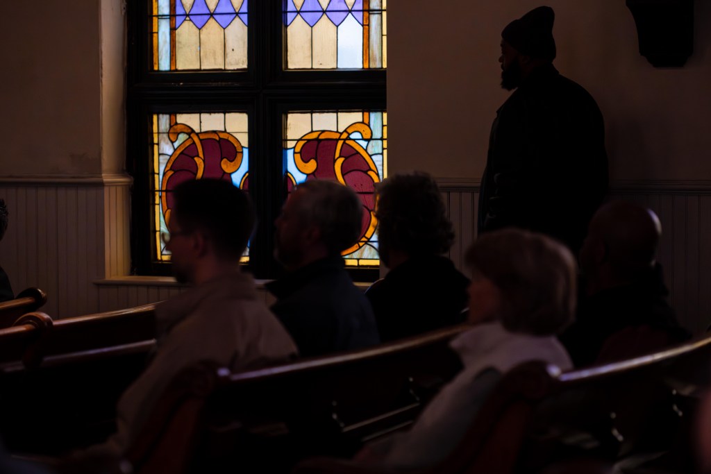 A photograph taken inside of a former church sanctuary. A few people are sitting in old wooden pews, and a tall man with a beard is standing nearby. Only their profiles are visible, but they're blurred out. Behind them, soft light filters in through stained glass windows.