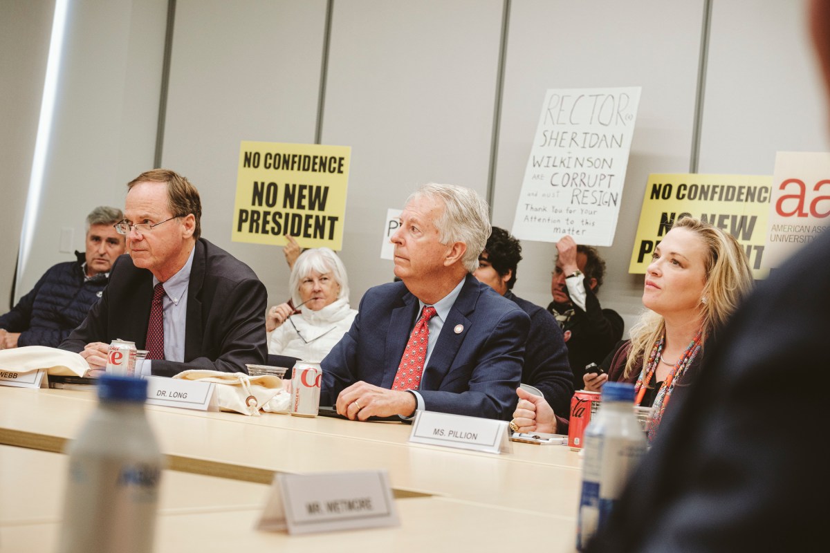 Two men and a woman in formal attire sit at a long table facing forward. Several people sit behind them holding up signs reading "No confidence, no new president," and "Rector Sheridan, Wilkinson are corrupt and should resign."