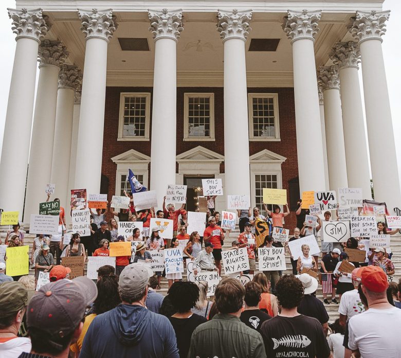 A crowd of people gather in front of a large brick building with white columns, holding up homemade signs.