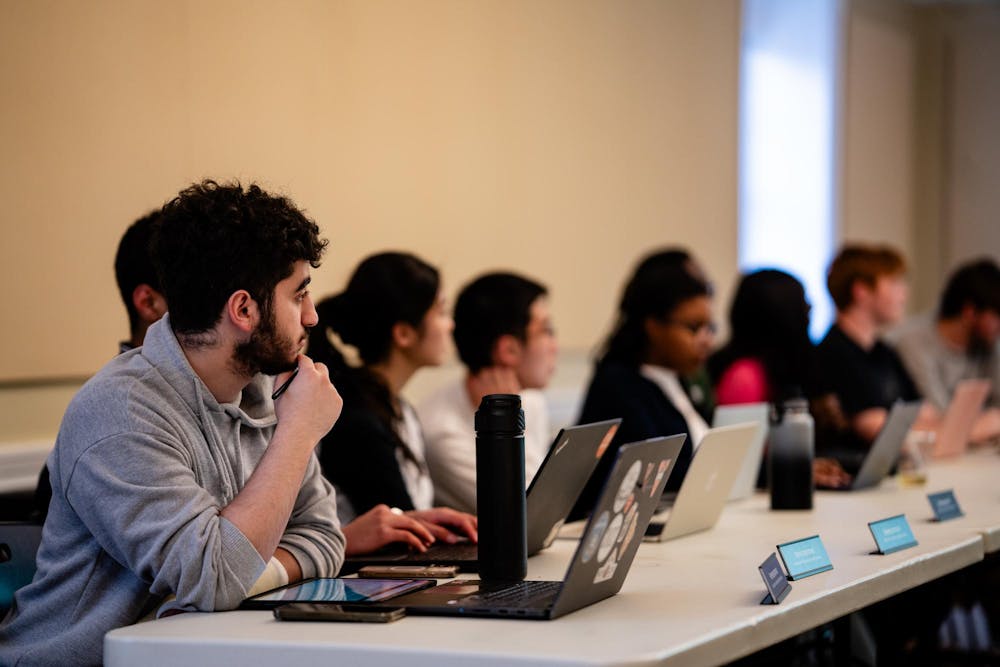 A row of people sit in front of laptops at a long grey table. The first man is in focus, the rest are blurred out.