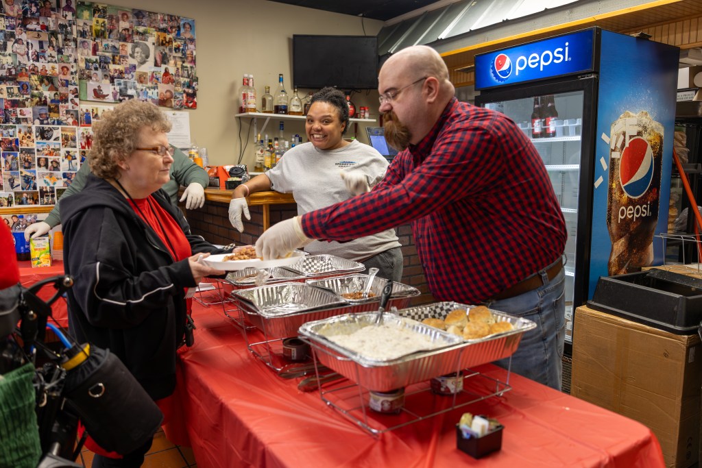A woman stands behind a table with several large trays of food, leaning on a nearby counter. A man stands behind the table and serves food onto a plate for a woman standing on the other side of the table. A drinks fridge and a wall covered with photos is visible behind them.