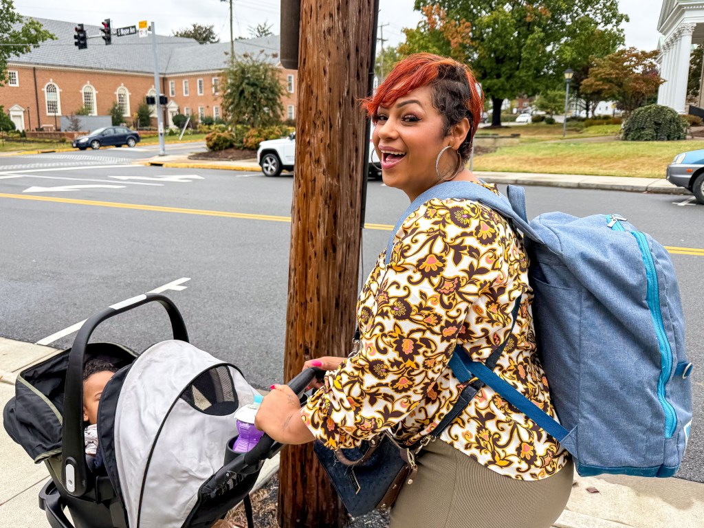 A woman pushes a stroller down the sidewalk and smiles at the camera. A street and brick buildings and a few cars parked by the side of the road are visible in the background.