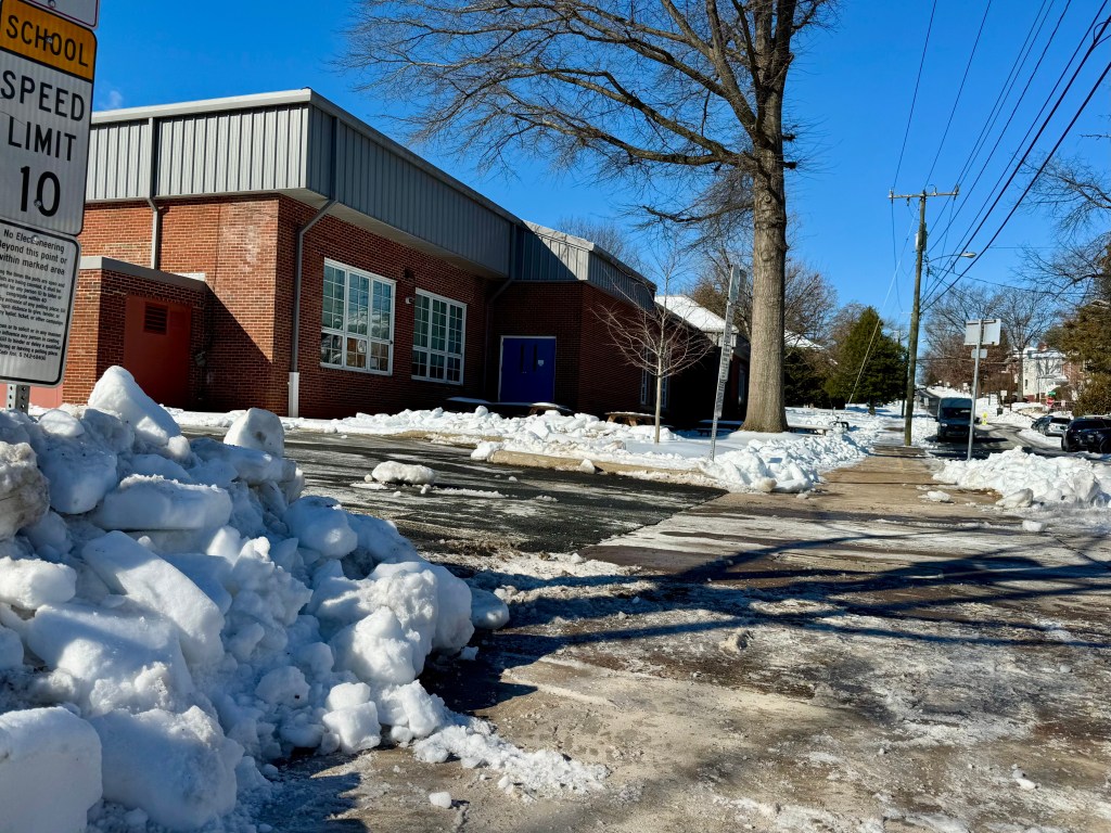 Sidewalks next to a brick building have just been cleared of snow, with snow and ice piled up next to them. Salt and snow litter the sidewalk.