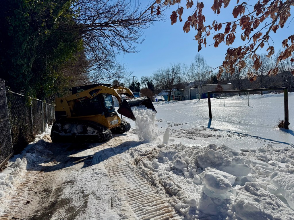 A bulldozer with tank treads drives down a small path near trees and a soccer field covered in snow and ice. The bulldozer is dumping snow onto the ground.