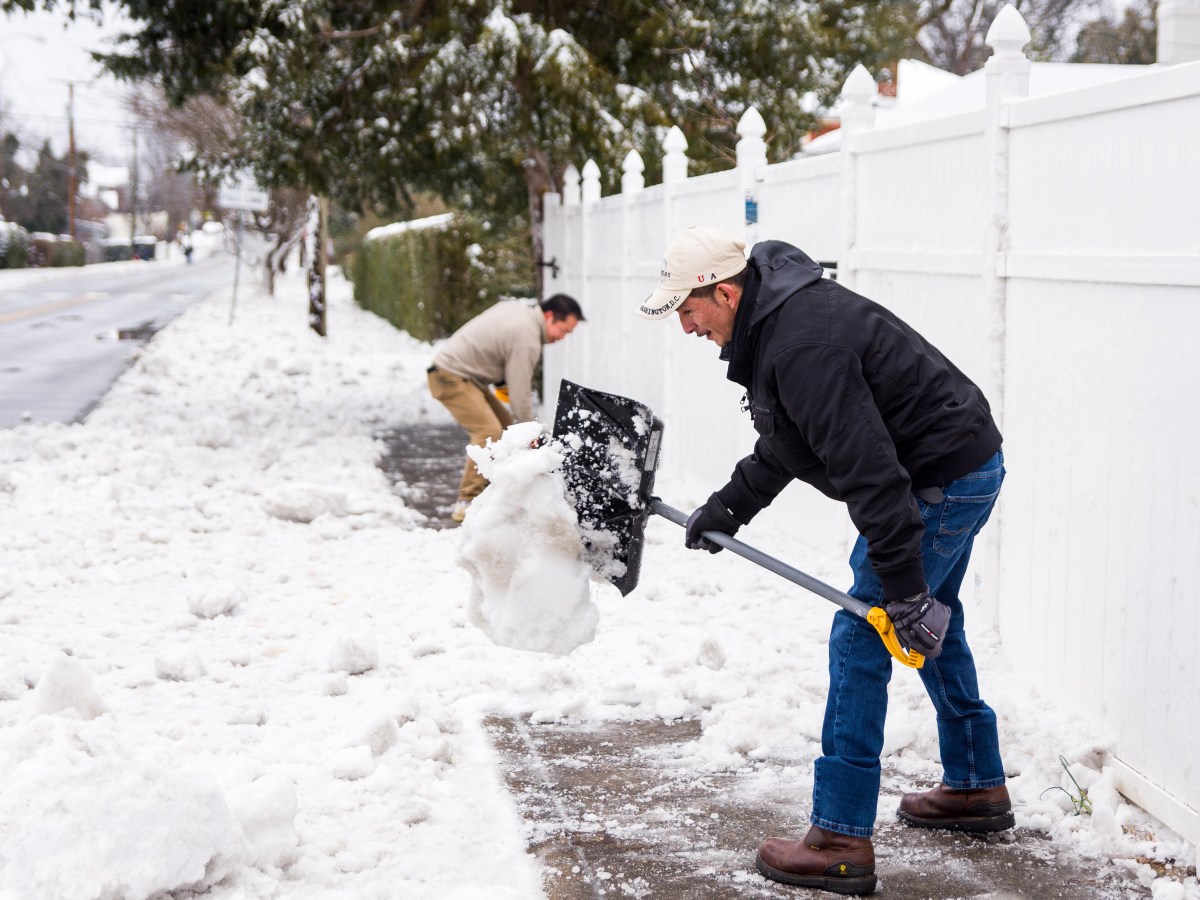 Two men with snow shovels clear a sidewalk next to a white fence. A pile of snow is visible off the curb beside them.