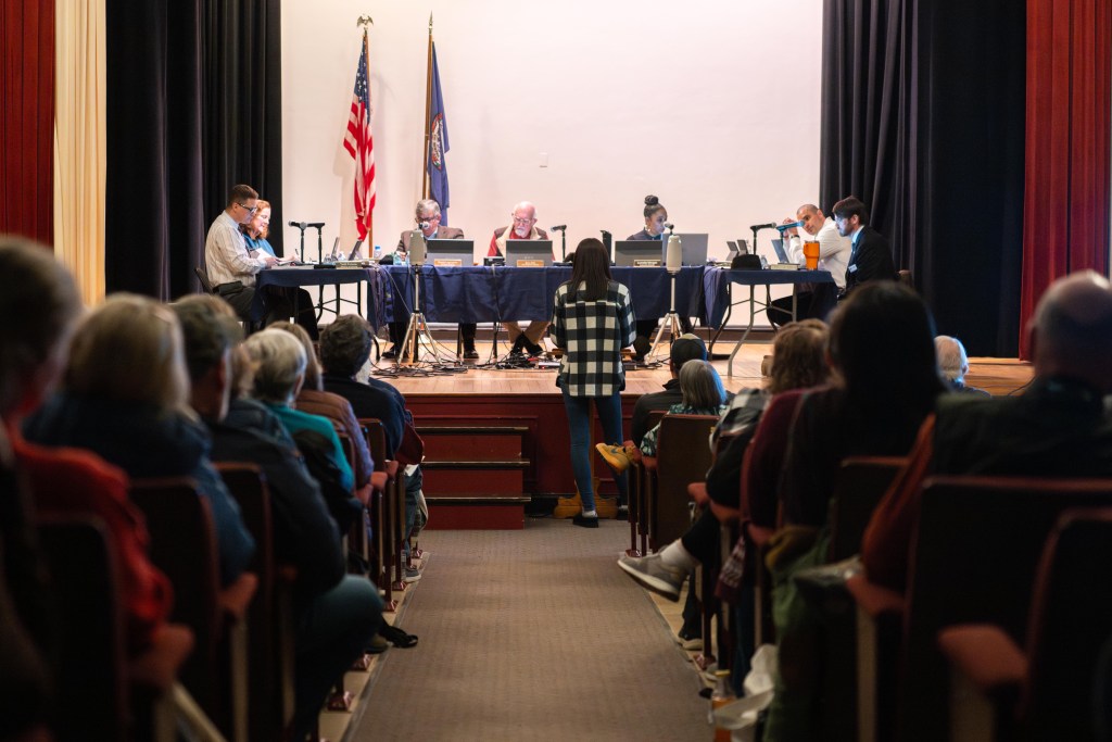Seven people sit at tables on a stage in front of dozens of others in an auditorium.