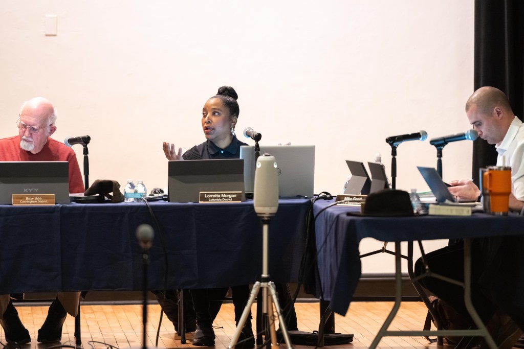 A woman speaks into a microphone. She is seated behind a table with others to her right and left.