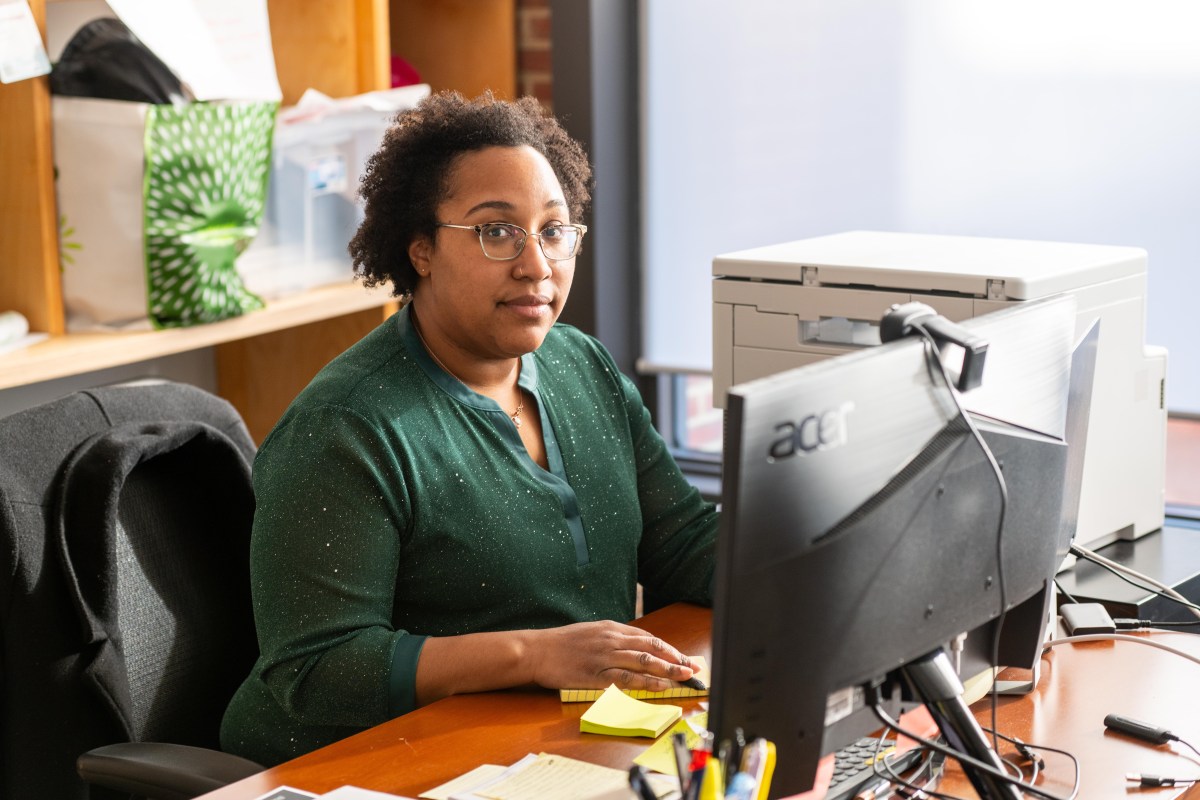 A Black woman sits at an office desk, in front of a computer screen. She is looking directly at the camera. There is a printer, some papers and a pad of sticky notes on the desk, as well as a cup full of pens and markers. On a shelf behind her are a shopping bag and a plastic bin full of supplies.