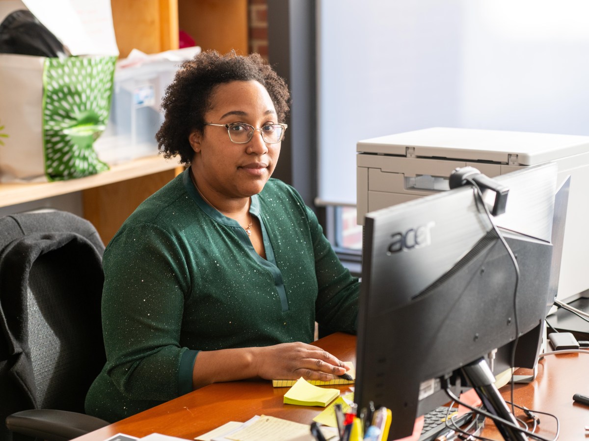 A Black woman sits at an office desk, in front of a computer screen. She is looking directly at the camera. There is a printer, some papers and a pad of sticky notes on the desk, as well as a cup full of pens and markers. On a shelf behind her are a shopping bag and a plastic bin full of supplies.