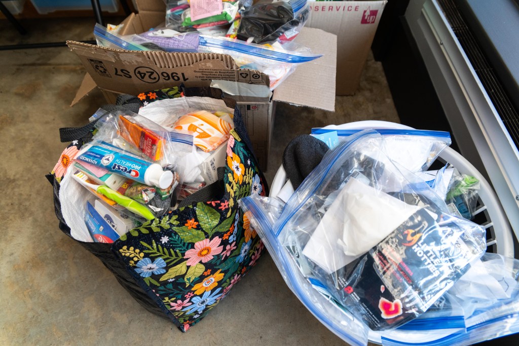 A photograph of supplies in an office. There are two large cardboard boxes, one large reusable grocery bag, and one plastic laundry basket. All of them are filled with plastic zip-top bags full of things like toothbrushes, toothpaste, hand-warmers, crackers, granola bars, bars of soap, wipes, and knit hats.
