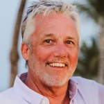 A man with white tousled hair smiles at the camera. He wears a light purple button-down opened at the collar.