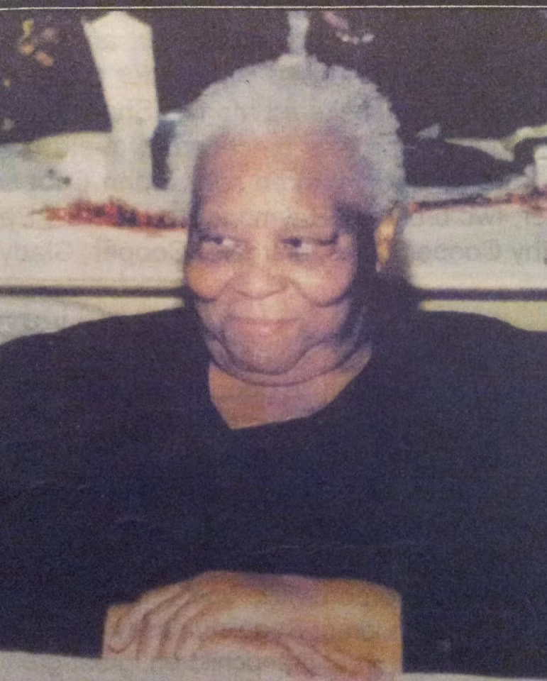 A woman with grey hair is seated with her hands resting on a table in front of her. She looks to the left and is smiling.