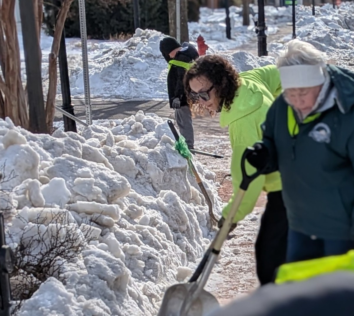 People in bright yellow coats shovel piles of snow along a sidewalk.