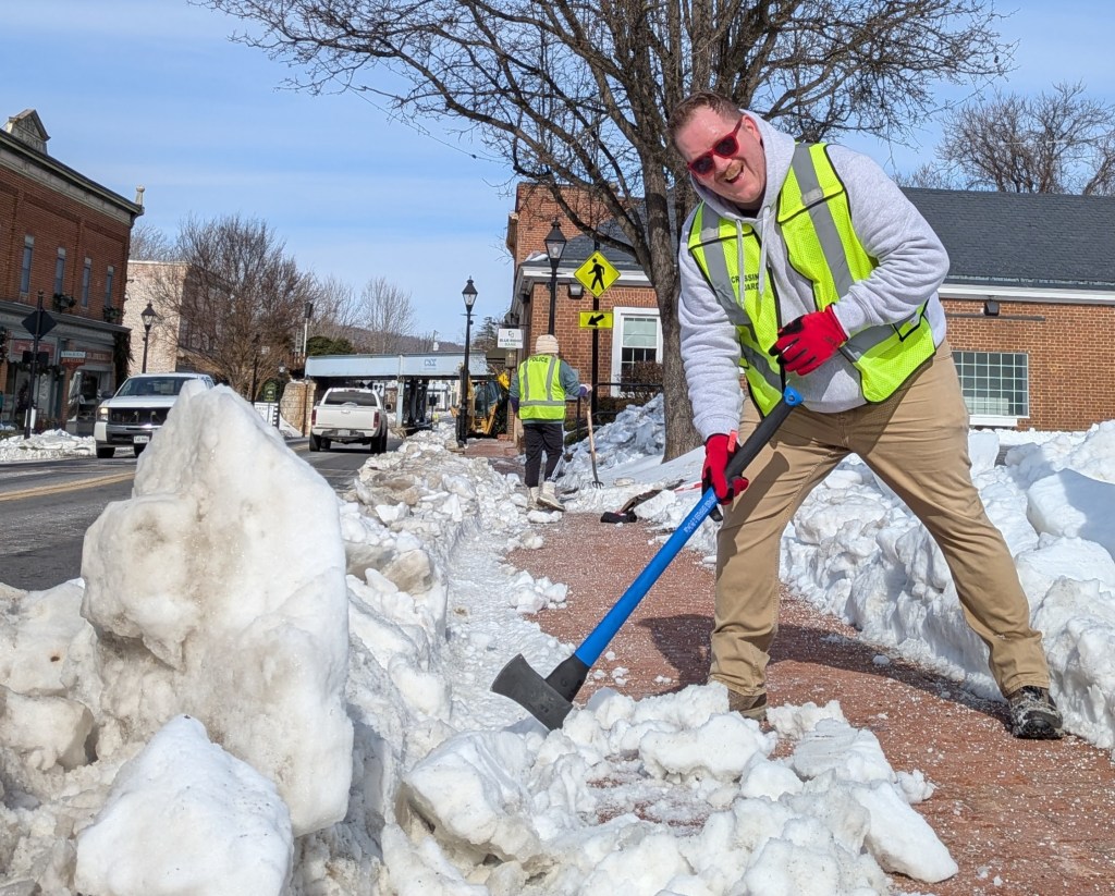 A man in a bright yellow safety vest and sun glasses poses with an ax by a pile of snow on a sidewalk.