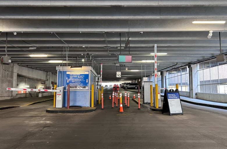 A photograph of the entrance/exit of a parking garage, taken in the daytime. There are three lanes, but the one in the center is blocked off by a collection of orange pylons and delineator posts with reflective tape at the top of each. A "Speed Limit: 10" sign hangs from the concrete ceiling. There is a booth for an attendant at the center-left of the photo. The left lane is for cars exiting, and the right lane is for cars entering. Two small cameras are attached to the ceiling, one above the right lane and one above the left.