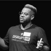 Black and white photo. Man against a black background speaking and his t-shirt reads "DisruptHR".