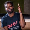 A man with short locs and a beard gestures with his hand. He wears a D.A.R.E TO RESIST DRUGS AND VIOLENCE shirt.