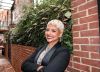 A woman wearing a blazer crosses her arms and smiles at the camera. She stand in front of a brick wall and foliage.