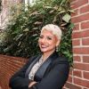 A woman wearing a blazer crosses her arms and smiles at the camera. She stand in front of a brick wall and foliage.