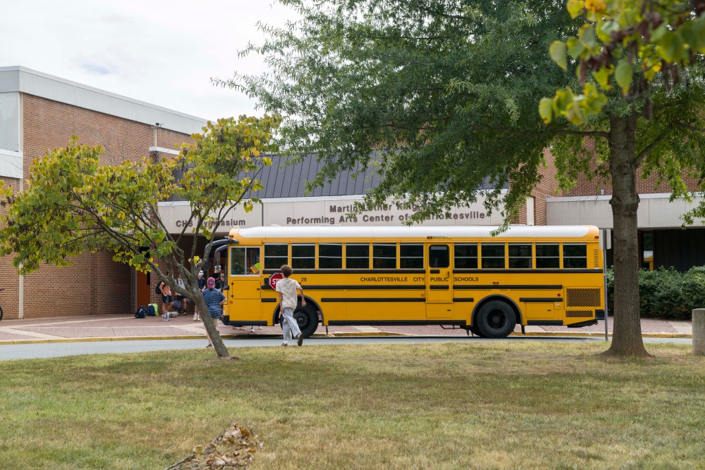 Students run through grass and rush past a yellow school bus toward Charlottesville High School.
