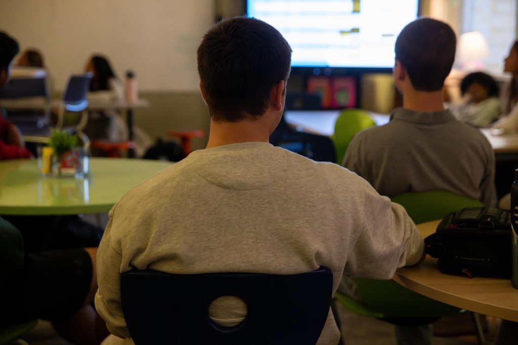 Several high school students are seated at round tables facing a large screen at the front of the classroom.