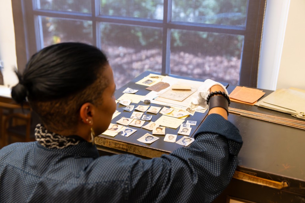 A photograph of a Black woman looking at archival materials laid out on top of a filing cabinet. The photo is taken from behind, so only the side of the woman's face is visible. She wears white archival gloves and holds a small rectangular photograph of a young child. The archival materials include various photographs of children as well as some written records.