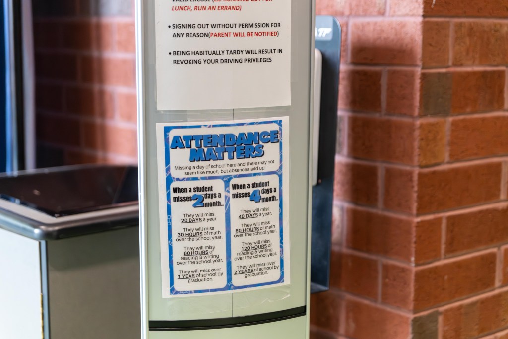 Signs posted next to a brick wall near a trash can and hand sanitizer dispenser. One sign reads "Attendance Matters" and says if students misses four days of school a month, they will miss over two years of school by the time they graduate.