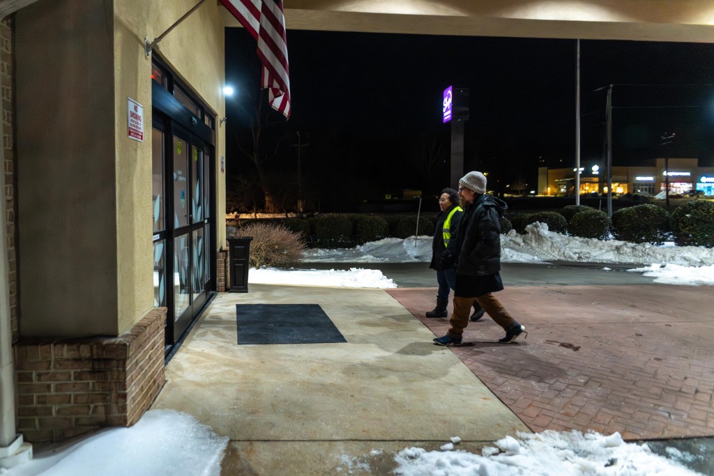 A photograph of two people walking side by side through the front door of a hotel. It is nighttime, and there are piles of snow and ice everywhere. One of the people wears a reflective vest, metal-rimmed glasses, a winter coat, long pants and boots. Another wears a knit cap, thick plastic glasses, a puffy winter coat, long pants, and sneakers with the laces untied.
