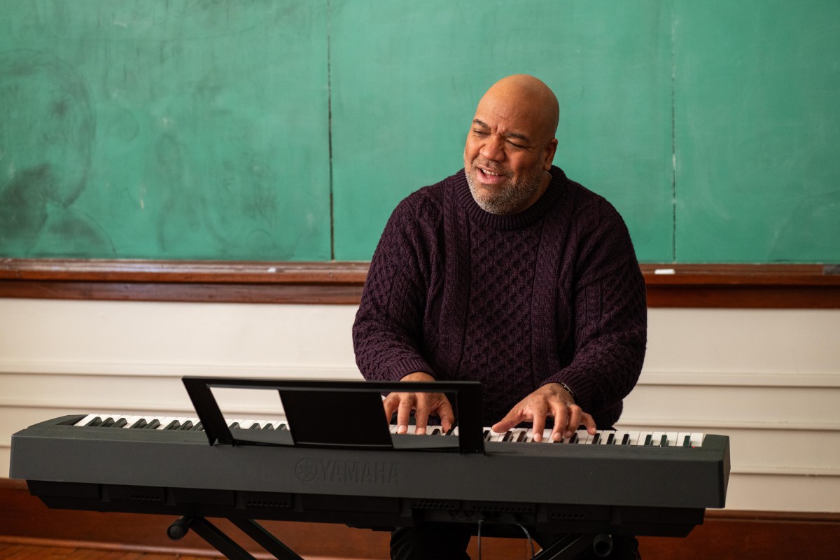 A photograph of a Black man sitting at a keyboard in a classroom, a wall of chalkboards behind him. He is playing the keyboard and singing.