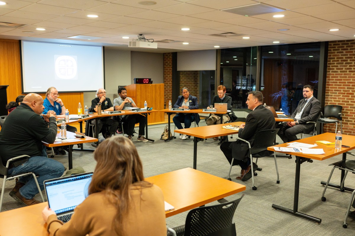 Several men and women are seated in a meeting room with brick walls at long tables forming a semicircle. At the center is another table where a man is seated, speaking and facing the other meeting participants.