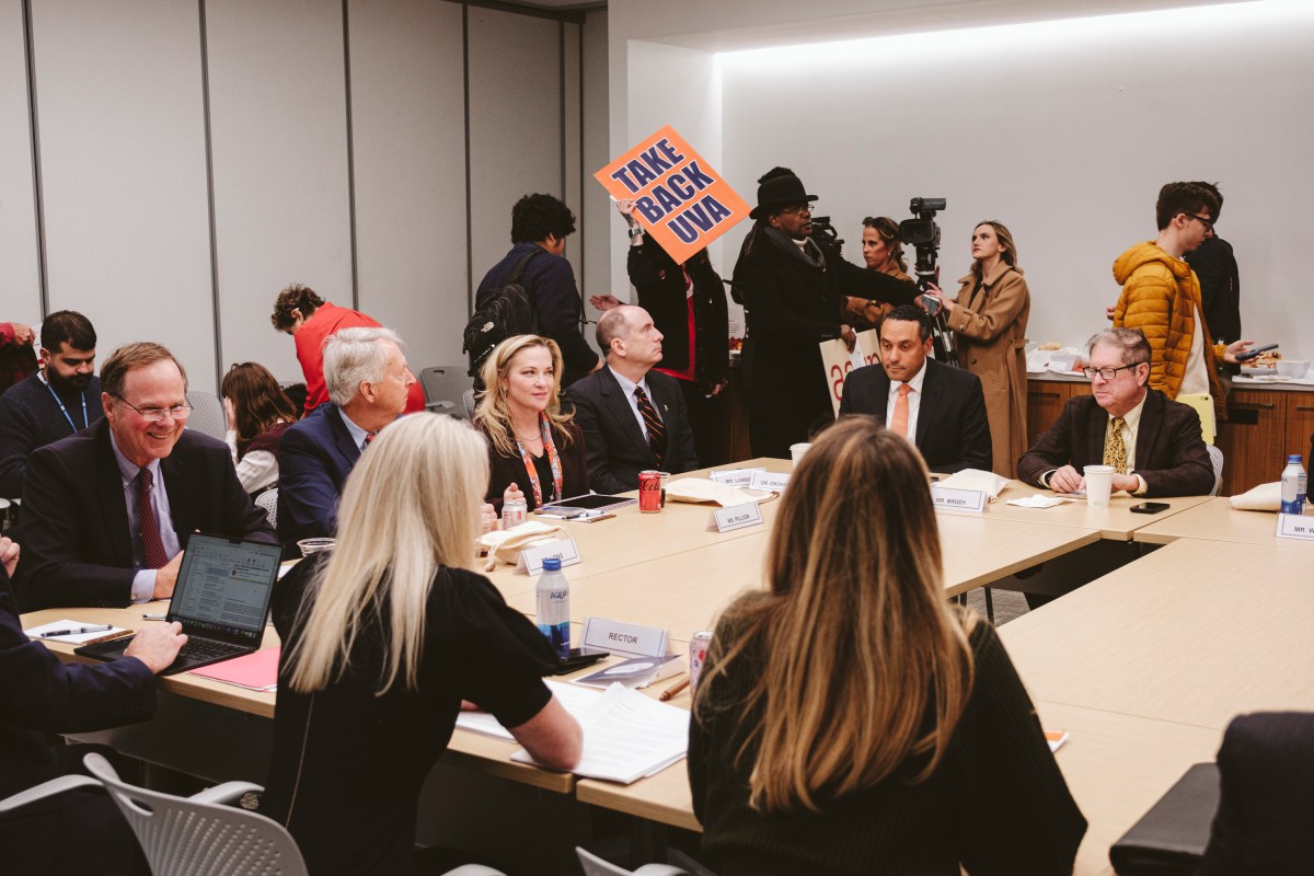 A group of men and women sit at a large conference table with name placards, notebooks, pens and drinks sitting in front of them. A small group of people stand and sit behind them with video cameras and signs reading "Take Back UVA."