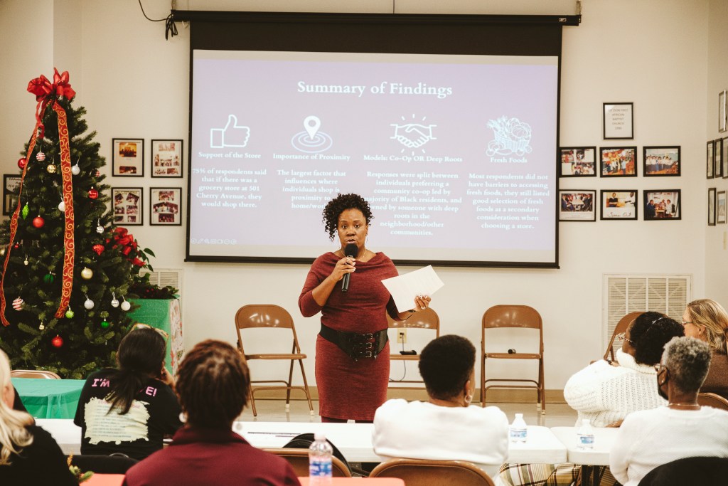 A photograph taken inside of a church hall during the Christmas season. A Black woman stands at the center of the photo with a microphone in one hand and a piece of paper in the other, talking to folks who have gathered in the room. Some of them are seated at a table in front of her, and only the backs of their heads and some of their profiles are visible in the photo.