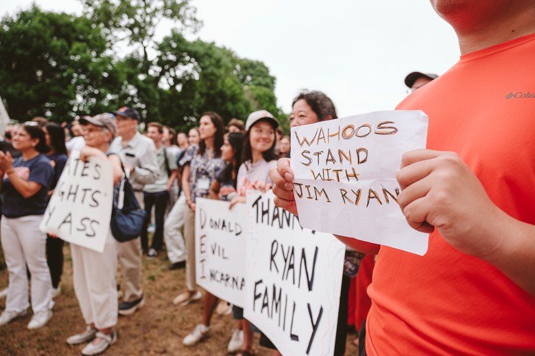 A group of protestors stand on dirt and grass near large trees, holding various homemade signs with various slogans, including "Wahoos stand with Jim Ryan."