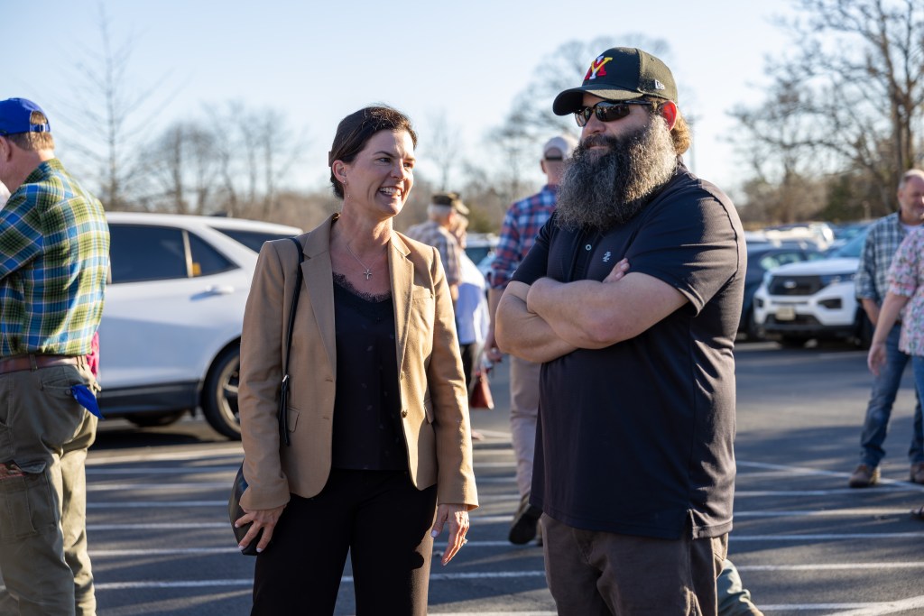 A woman with brown hair wearing a tan jacket stands in a parking lot while speaking with a man with a large beard, sunglasses and a dark baseball cap.