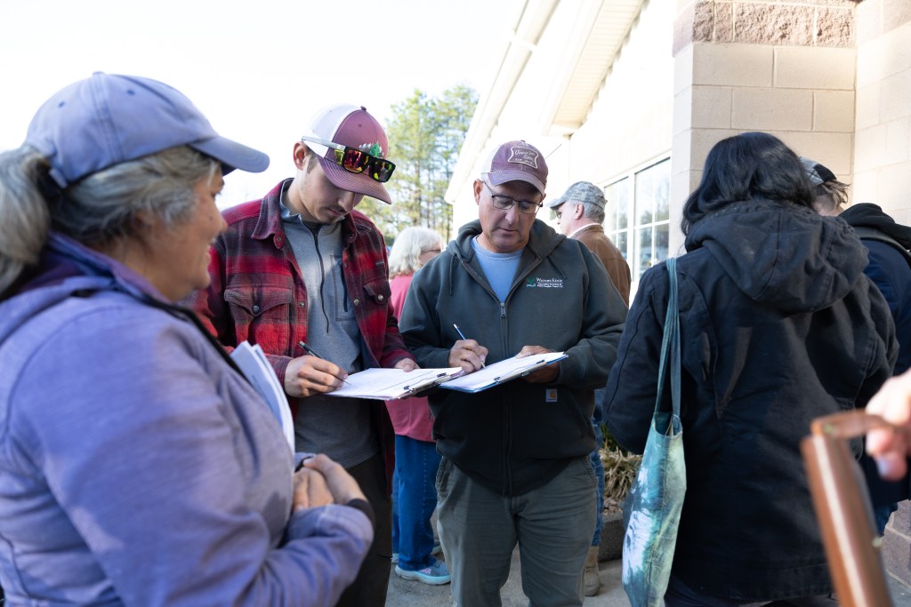 A woman in a purple coat and hat stands at left as two men in casual clothing and baseball caps sign petitions on clipboards. A beige building is visible in the background.