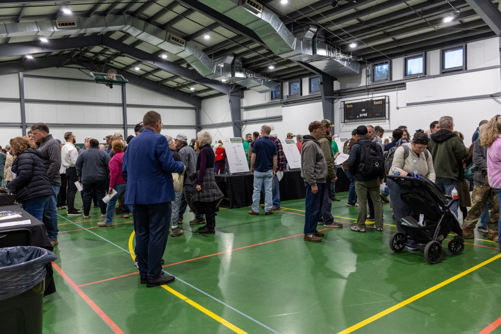 A large crowd stands inside of a gymnasium with a green floor. Several people can be seen looking at white posters.