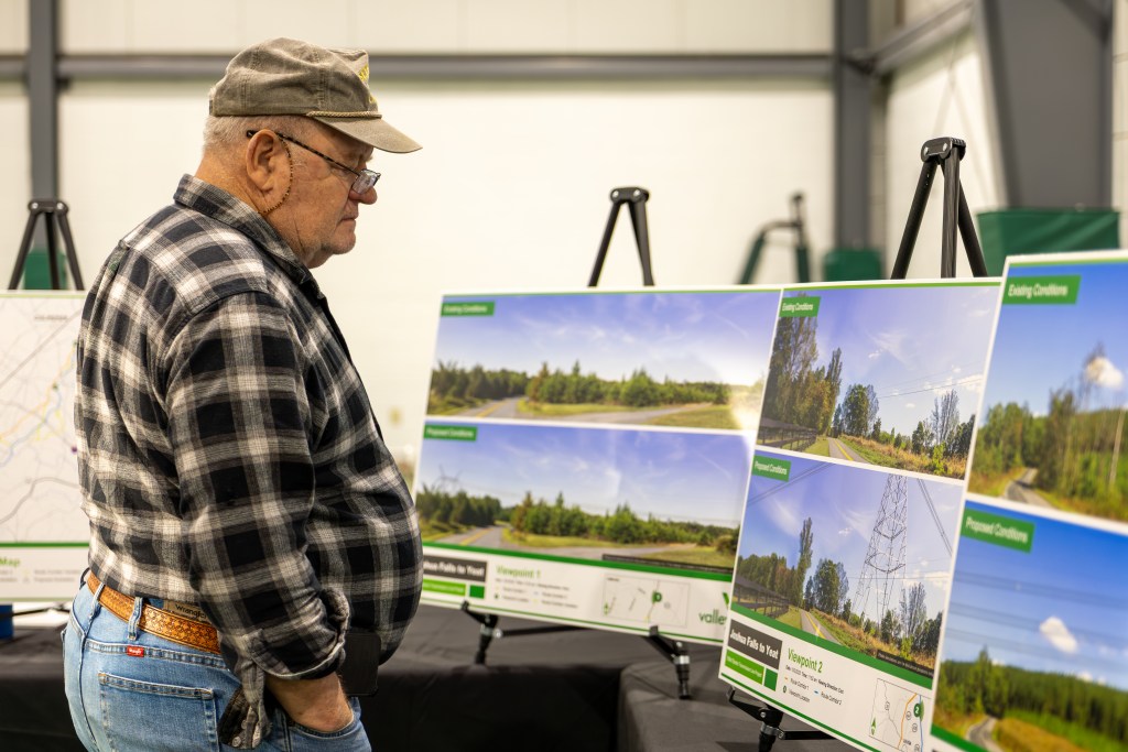A man in a tan hat and plaid shirt looks at several posters showing rural landscapes with transmission lines.