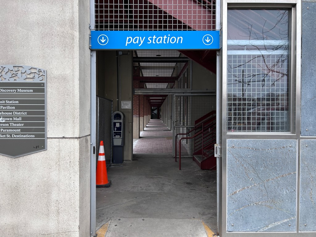 The ground floor of a parking garage. At the center is an open doorway, with a concrete wall to the left and a metal and glass wall to the right. Over the doorway is a sign that reads "pay station" with two arrows pointing downward. Beyond the doorway are metal and concrete stairs, and, to the left, a payment kiosk. A chain-link fence is pushed up close to the kiosk and perpendicular to it, and there is a traffic cone in front of the fence.