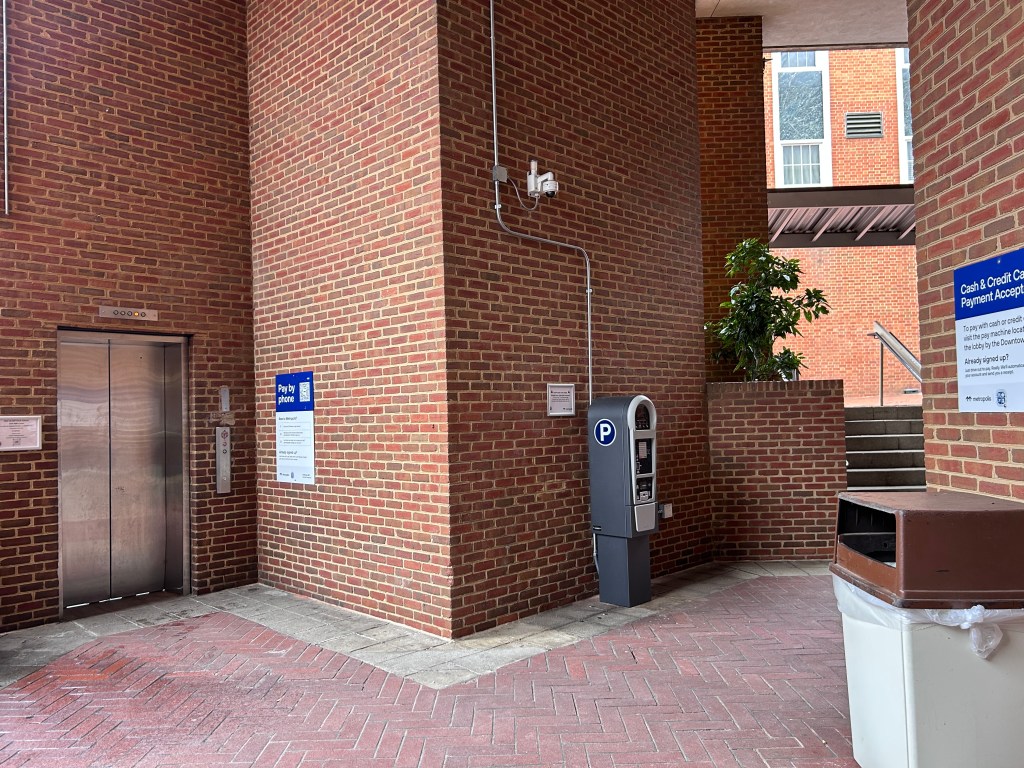 A ground-level brick structure on a brick-paved floor. At the center of the photo is a payment kiosk. To the right of the kiosk is an elevator door. To the left of the photo, there is a trash can. There are also two signs with instructions for how to pay for parking.