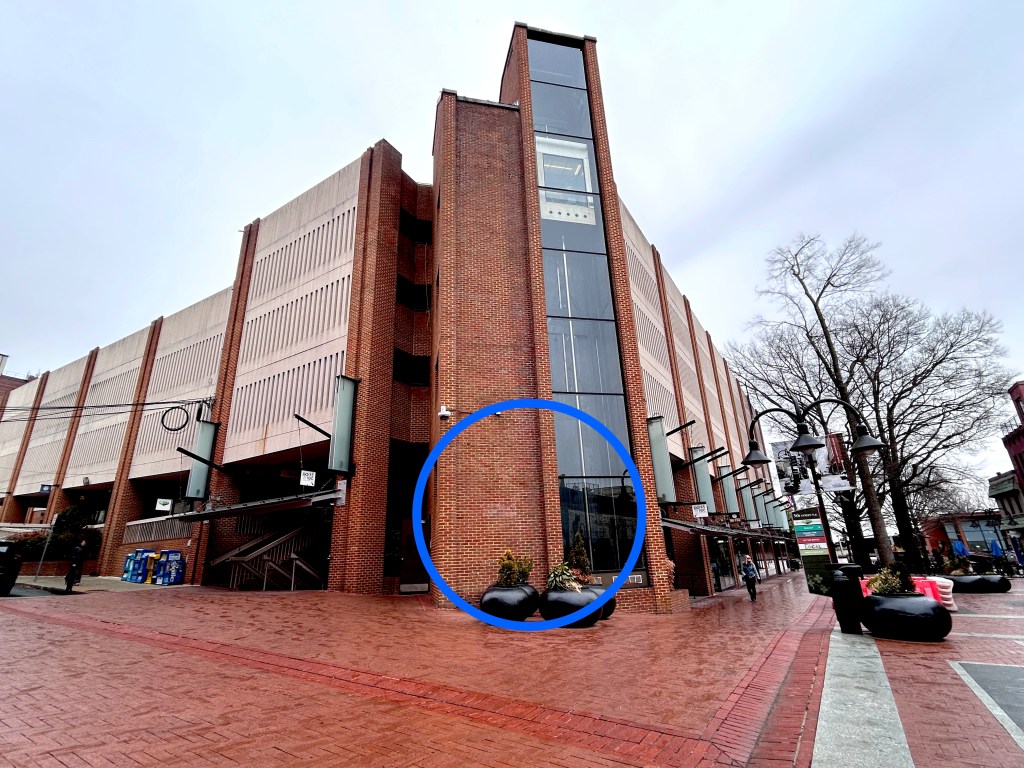 A four-story brick and concrete building situated on a brick-paved street on a rainy day. The bricks are wet, and there are bare trees and lampposts, as well as large oval planters, visible to the right side of the building. There is a glass elevator and a brick column at the center of the photo. A drawn-on circle is meant to show the specific location of a payment kiosk.