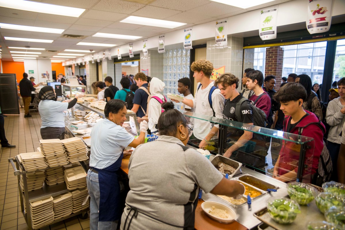 Several people stand behind a counter preparing and serving food as a long line of high school students stretches down the counter and into the hallway.
