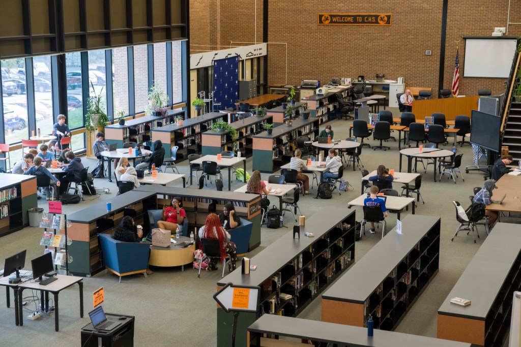 A large open space with several book shelves, desks, long conference tables, and stands with computers. High school students are walking around the room and are seated at tables, reading and looking at papers or laptops.
