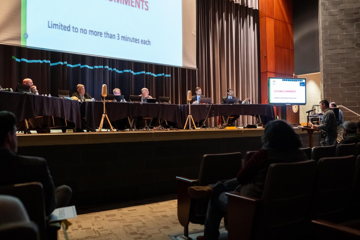 In a dark auditorium, several men sit at long tables set up on stage. Several people sit in rows of chairs facing the stage, and a man stands at a pedestal with a microphone facing the people on stage.