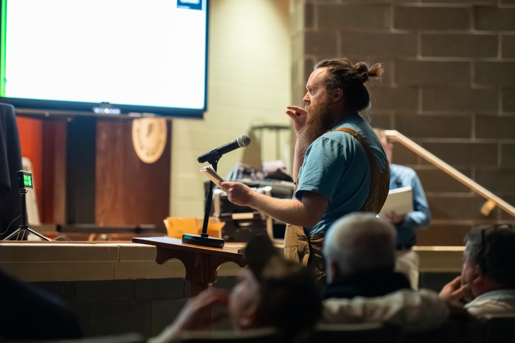 A man stands at a podium, speaking into a microphone while holding a stack of paper in one hand and gesturing with his other hand.