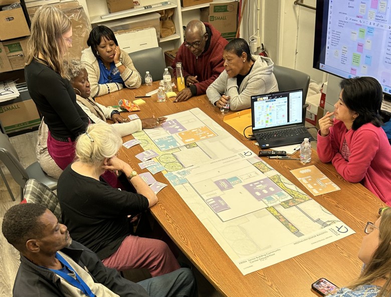 A photograph of a racially diverse group of nine people sitting in a room around a long table. They're all looking thoughtfully at an architectural drawing printed out on a long sheet of paper and placing smaller squares and rectangles of paper on top of it. Those smaller papers represent different parts of the planned development, such as residential buildings, parks and community spaces.